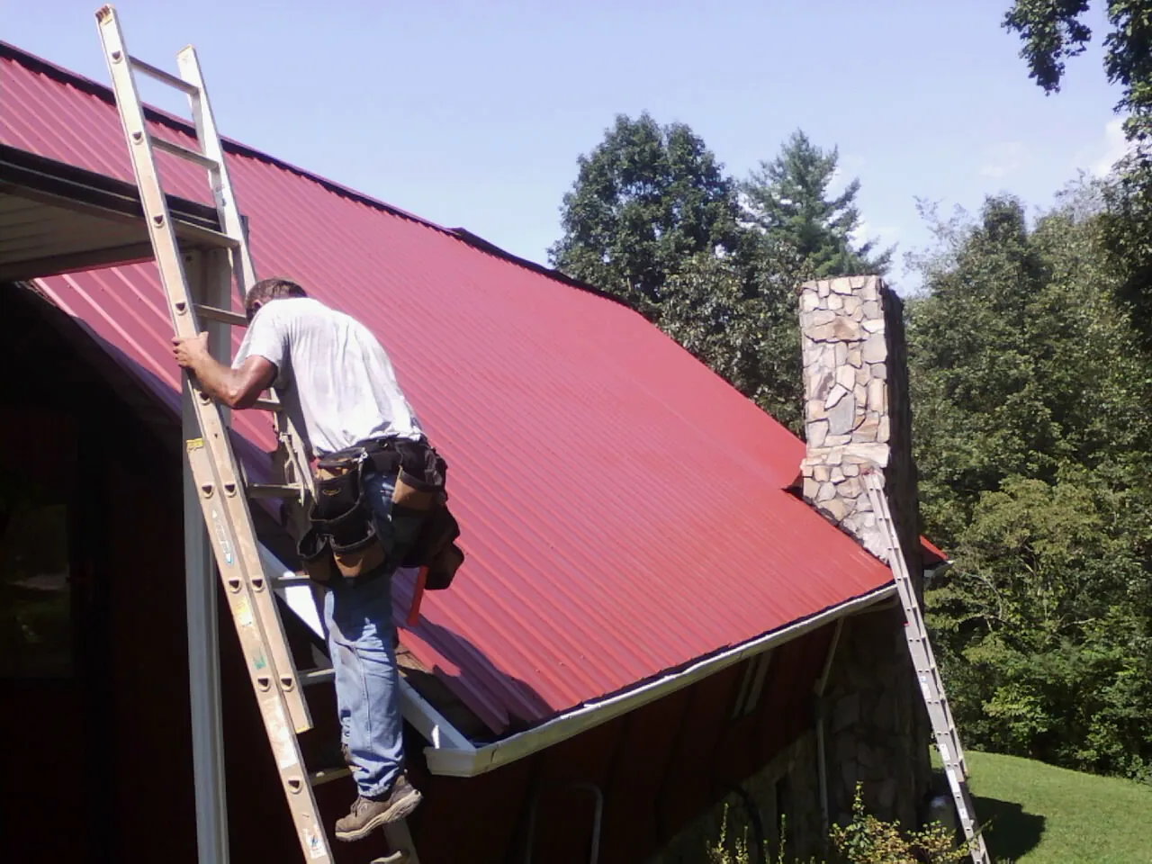 Roofer on a ladder beside a red metal roof and stone chimney — Soffit & Fascia Repair by Roofing Brothers in Hawthorne